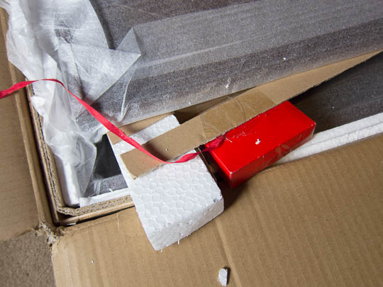 The small, red cardboard box is attached to packaging elements inside the large carton. The red ribbon attaches to that small box.