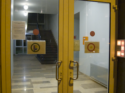 Double-winged glass doorway shown from the outside with a button panel at the right. Both doors have identical handles that are located in the center of the doorway, and the right door's hinges are visible. A circular sign is posted on either door.