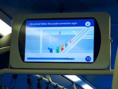 A monitor suspended from the bus' roof displays comic-style people on an escalator and states,'On arrival follow the purple connection signs'. The progress bar at the bottom shows that the bus has already gone three quarter of the way to its destination, Terminal five.