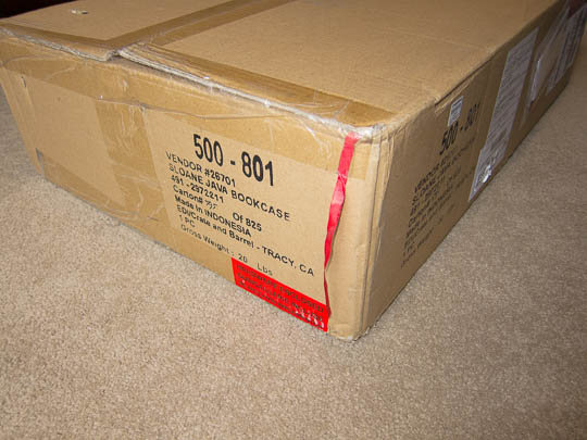 The large carton containing the shelves' parts. The red ribbon is clearly visible on the cardboard.